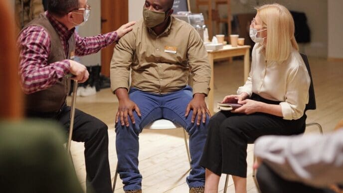 Two men in casual clothing and face masks comfort each other during a mental health support group session.