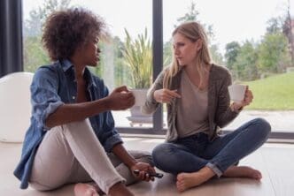 Two smiling young women of diverse ethnicities sit on the floor by a window, drinking coffee and using a smartphone in a luxurious home.