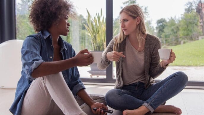 Two smiling young women of diverse ethnicities sit on the floor by a window, drinking coffee and using a smartphone in a luxurious home.