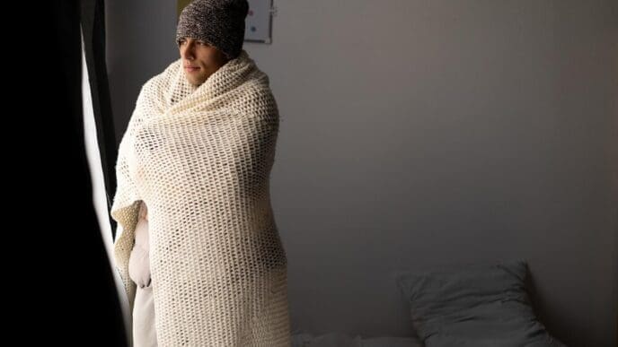 A man wrapped in a blanket sits in a cold bedroom, likely suffering from the lack of heat.