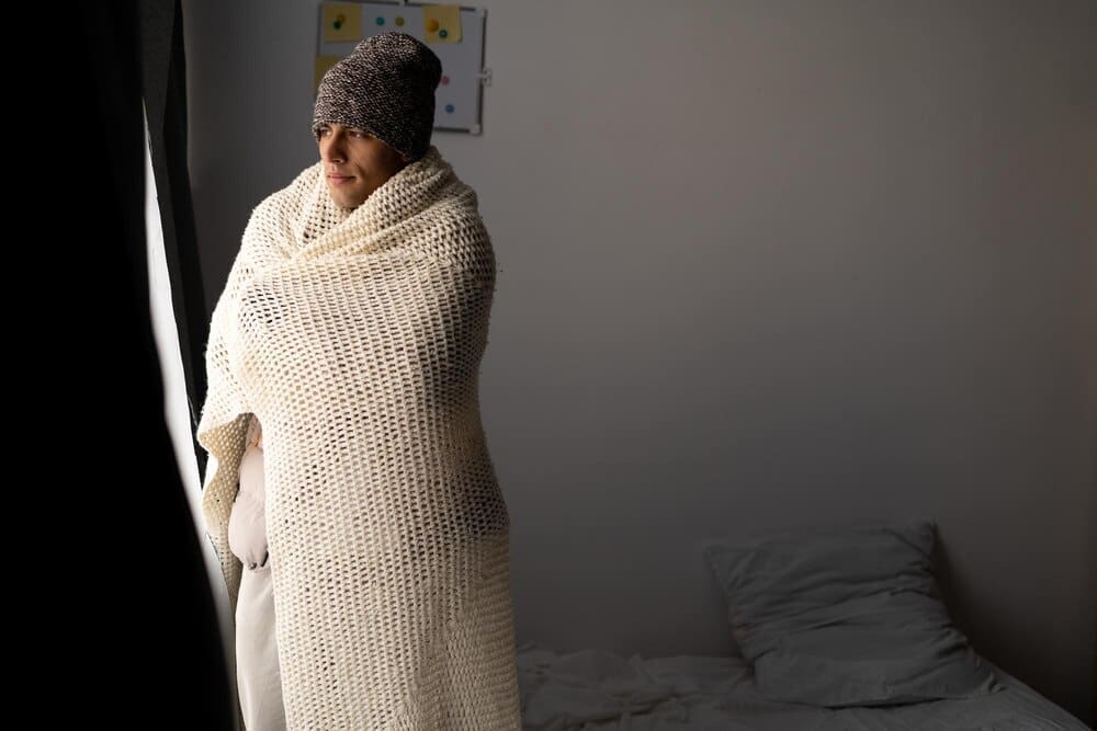 A man wrapped in a blanket sits in a cold bedroom, likely suffering from the lack of heat.