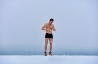 A shirtless man stands in the snow, presumably for a winter health practice.