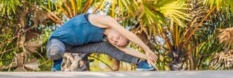 Woman in athletic wear performs a yoga pose on a mat in a park with lush, tropical foliage.