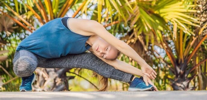 Woman in athletic wear performs a yoga pose on a mat in a park with lush, tropical foliage.