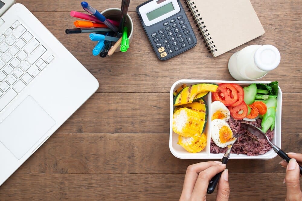 Woman at a desk enjoys a healthy breakfast from a box containing rice berry and a boiled egg.
