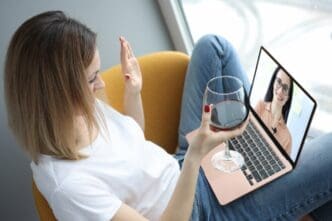 A woman smiles and waves at her friend on a video call while holding a glass of wine.