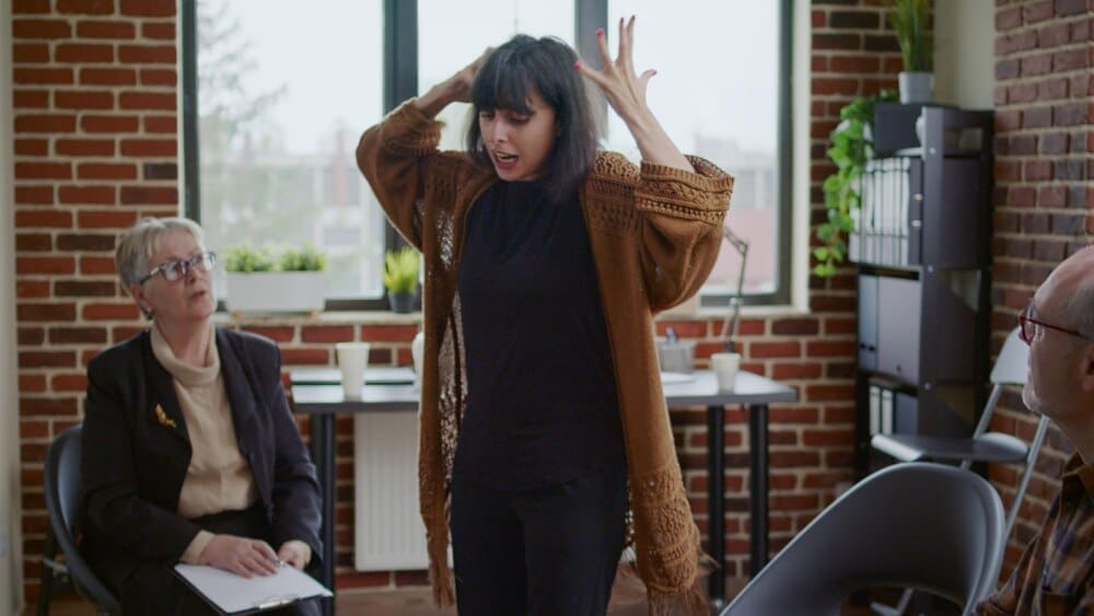 A woman with mental health issues has an angry outburst during a therapy session, observed by a group of people and a psychologist.