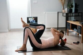 Woman in workout clothes using a laptop to follow along with a fitness trainer during a remote exercise class in her living room.
