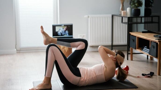 Woman in workout clothes using a laptop to follow along with a fitness trainer during a remote exercise class in her living room.