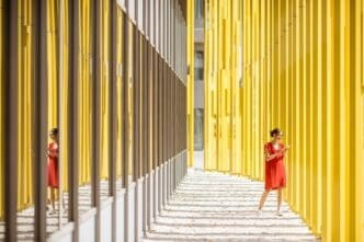 A woman in a red dress stands in front of a modern yellow building, part of an abstract geometric composition.