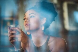 A young woman sips a cappuccino while looking out a window, lost in thought at a coffee shop.