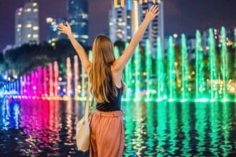 A woman stands in front of a colorful fountain on a lake at night with the Petronas Twin Towers and the Kuala Lumpur skyline in the background.