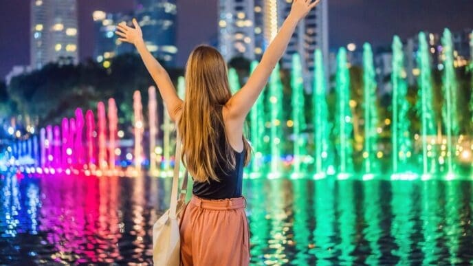 A woman stands in front of a colorful fountain on a lake at night with the Petronas Twin Towers and the Kuala Lumpur skyline in the background.