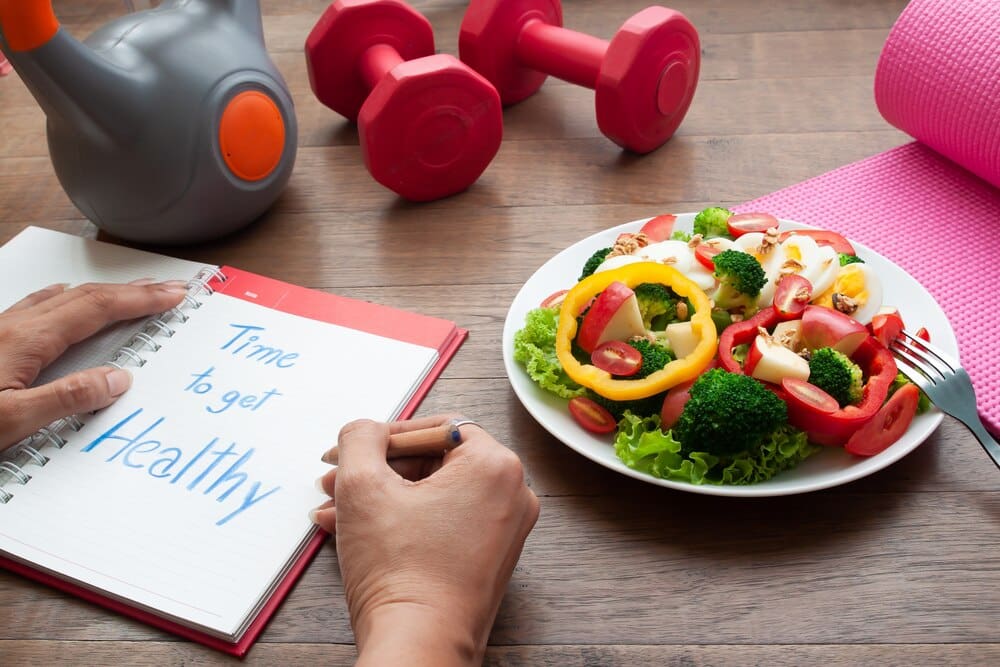 A woman writes in a diary on a table with a salad and fitness equipment.