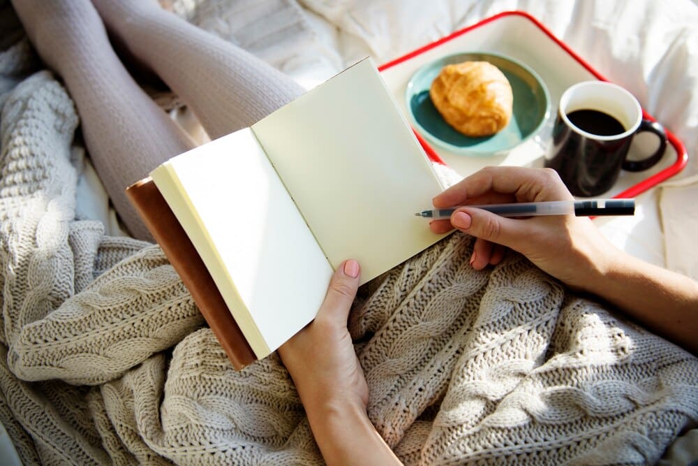 A woman sits on a bed in the morning, writing in a notebook with breakfast nearby.