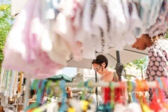 Two women browse clothing displayed on racks at an outdoor market.