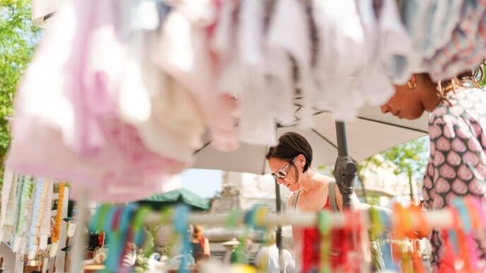 Two women browse clothing displayed on racks at an outdoor market.