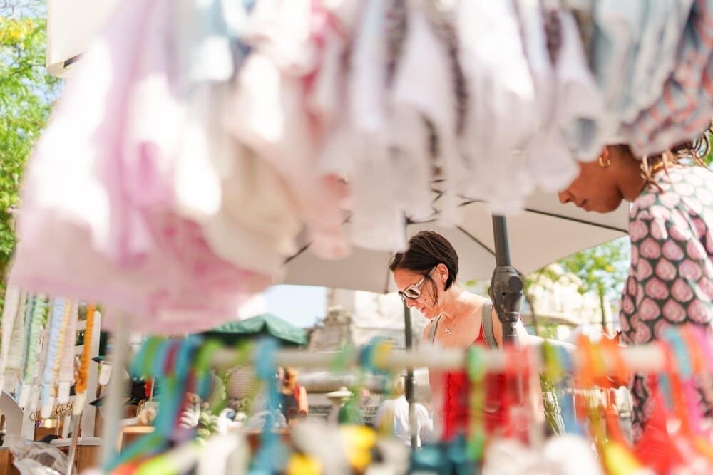 Two women browse clothing displayed on racks at an outdoor market.