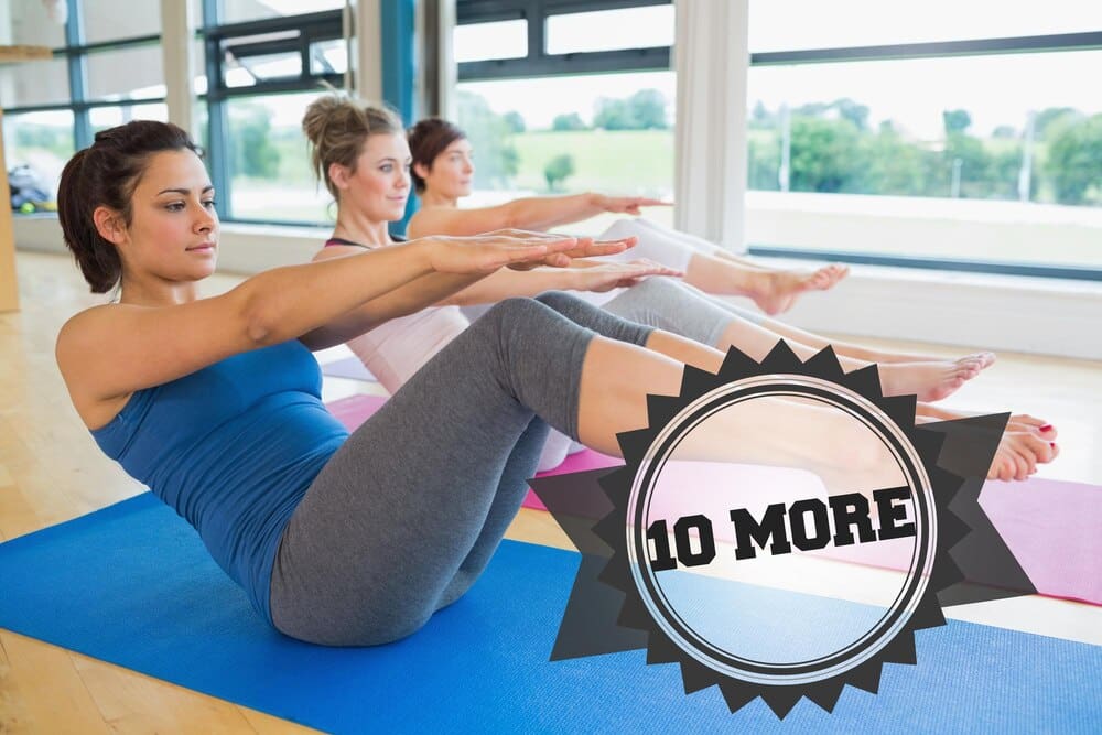 A group of women practice the boat pose in a yoga class, with a badge visible in the background.