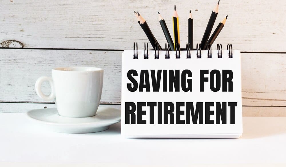 A white notepad with "SAVING FOR RETIREMENT" written on it sits next to a white cup of coffee on a light surface.