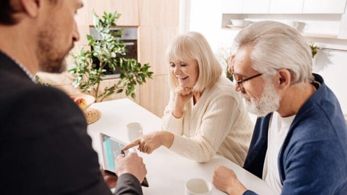 A real estate agent assists a retired couple, pointing at a project on a tablet.