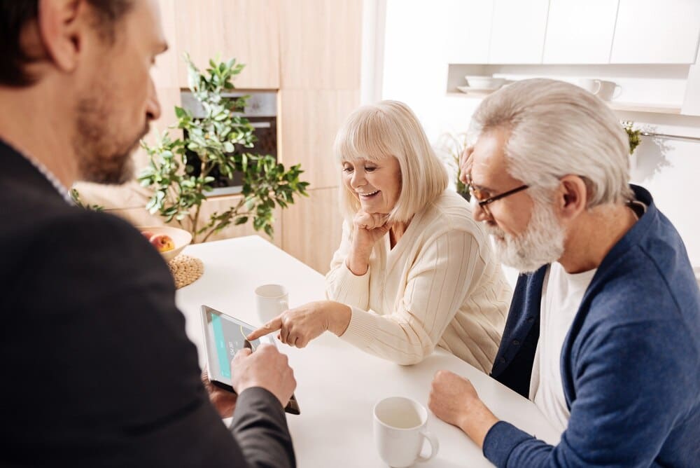 A real estate agent assists a retired couple, pointing at a project on a tablet.