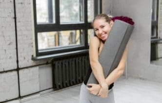 A smiling woman holds a yoga mat in a studio, preparing to work out.