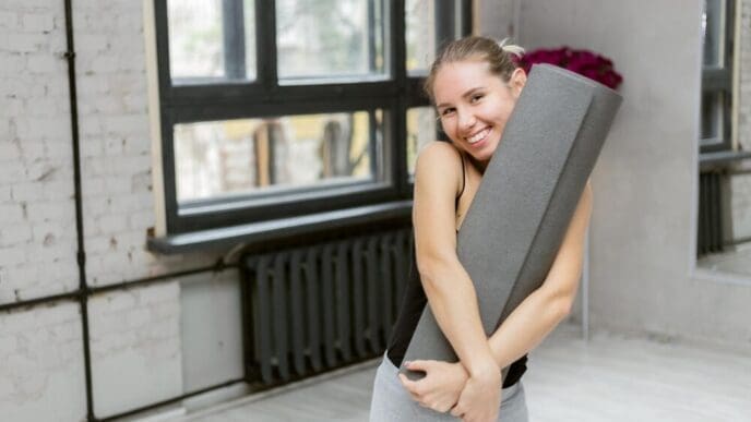 A smiling woman holds a yoga mat in a studio, preparing to work out.