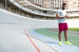A young man with a beard wearing running clothes checks his fitness tracker in front of a tall building.