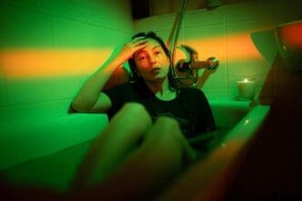 A young Asian woman in a t-shirt sits in a bathtub under a neon light, conveying a sense of depression.