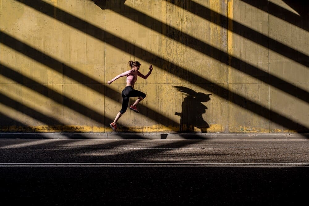 A young, blonde woman in athletic clothing jumps in the middle of a street.
