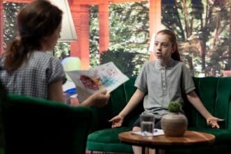 A young girl with a furrowed brow and clenched fists appears angry while reviewing disturbing drawings with a psychologist.