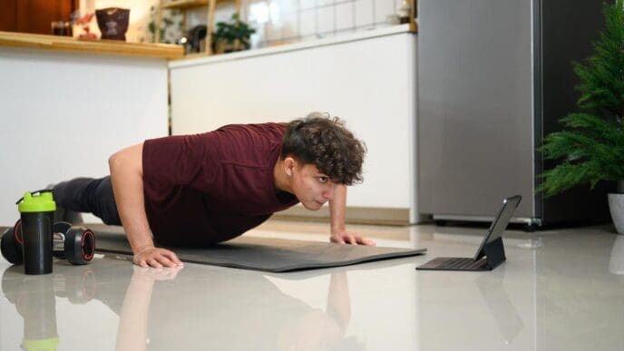 Young man in workout clothes doing push-ups while watching a fitness video on a tablet.