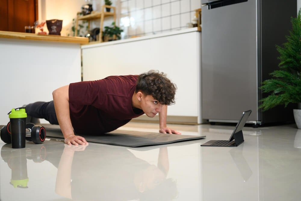 Young man in workout clothes doing push-ups while watching a fitness video on a tablet.