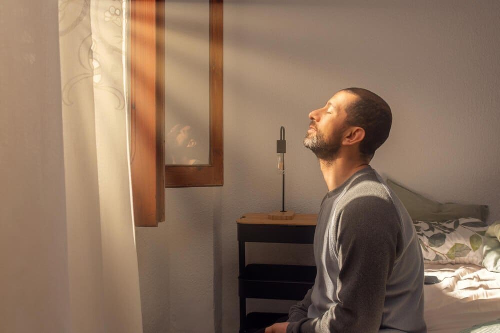A young man sits on his bed, bathed in the morning sunlight streaming through his window.