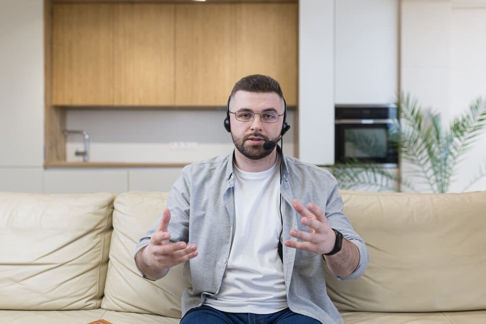 Young man wearing a headset smiles while using video chat at his desk in a home office.