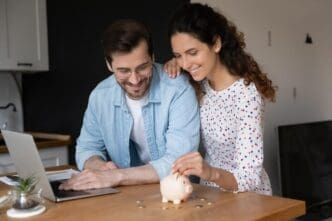 A young couple in their kitchen puts coins into a piggy bank while using a laptop, symbolizing financial planning and saving for a down payment on a house.