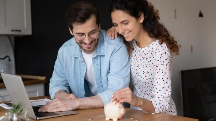 A young couple in their kitchen puts coins into a piggy bank while using a laptop, symbolizing financial planning and saving for a down payment on a house.
