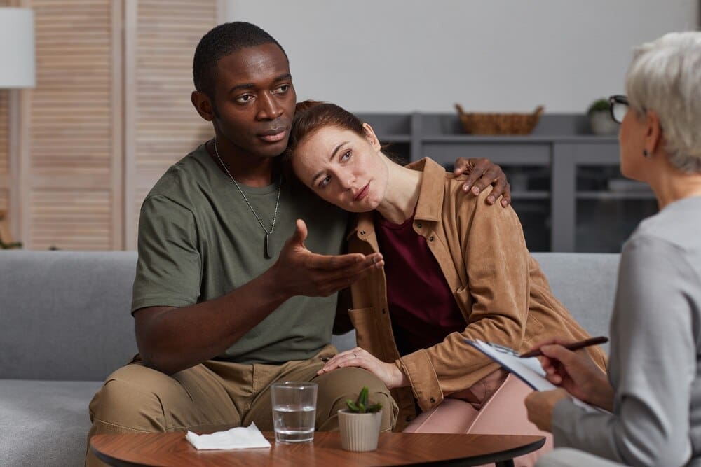 A young multiethnic couple sits together, supporting each other during a consultation with a psychologist.