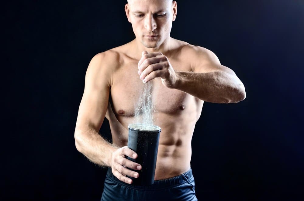 A young, muscular man pours protein powder from a container into a black shaker bottle against a dark studio backdrop.