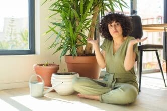 A young woman who appears to be of Arab descent sits cross-legged on the floor next to several potted plants and a watering can, shrugging with a confused or uncertain expression while looking directly at the camera.