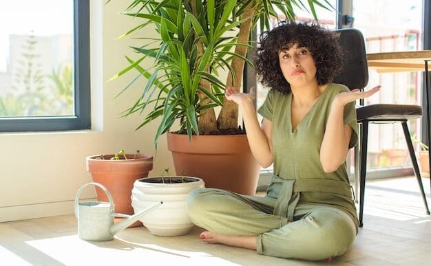 A young woman who appears to be of Arab descent sits cross-legged on the floor next to several potted plants and a watering can, shrugging with a confused or uncertain expression while looking directly at the camera.