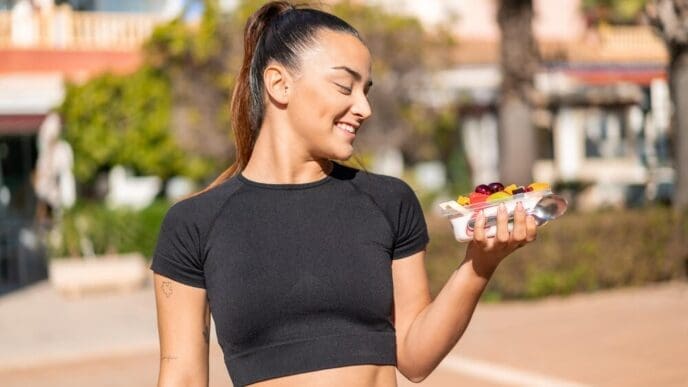 A young, attractive brunette woman holds a bowl of fruit while standing outside.