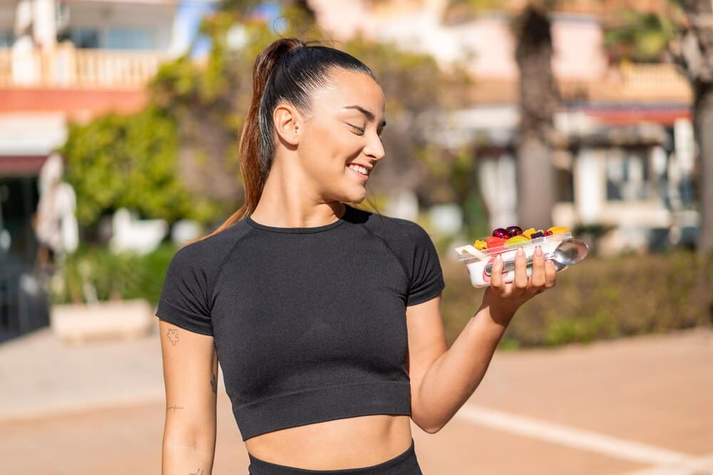 A young, attractive brunette woman holds a bowl of fruit while standing outside.