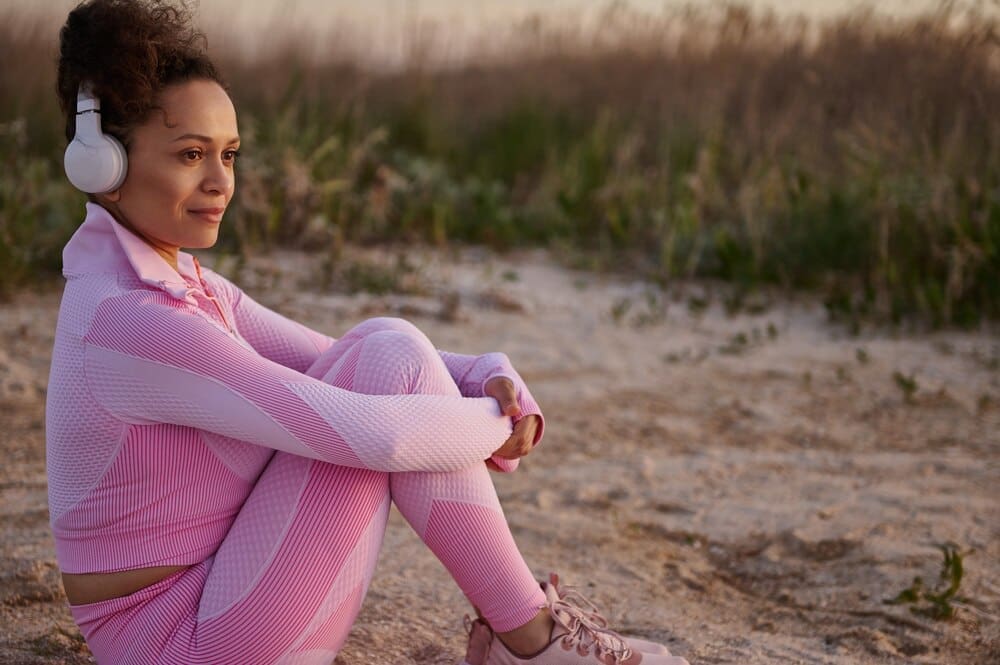 Young woman in pink sportswear sits on a riverbank, enjoying nature.