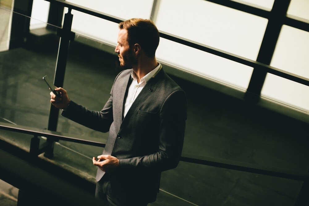 Young businessman in a grey suit stands near a modern building, holding a smartphone.