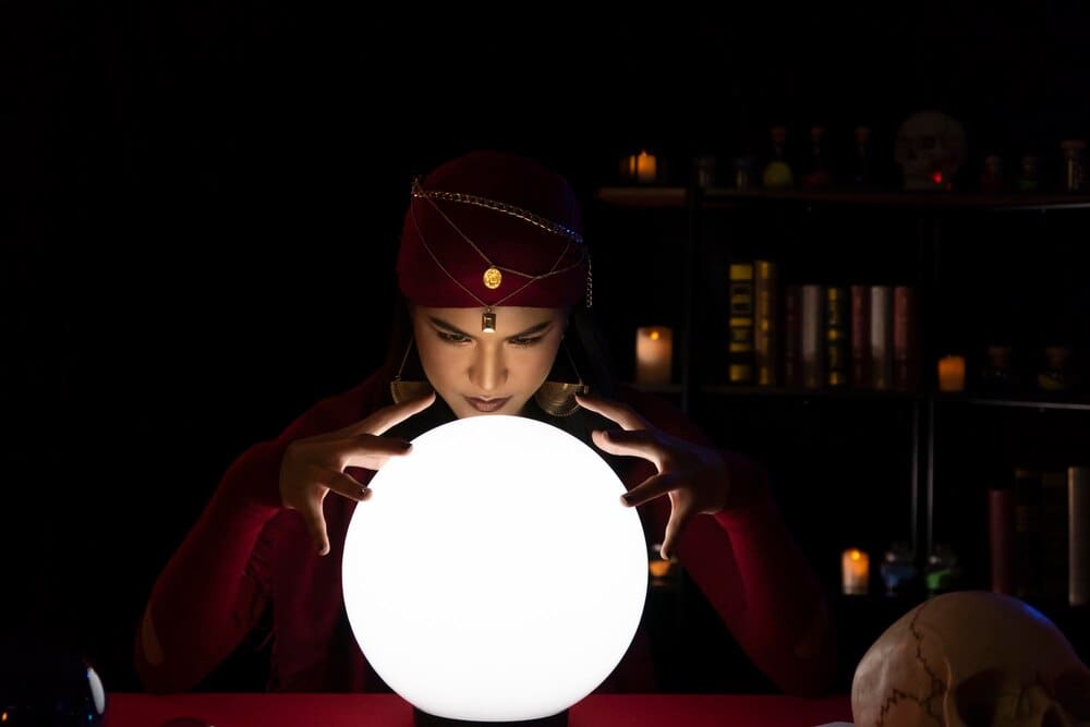 A young woman gestures towards a glowing crystal ball in a dimly lit room.