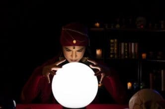 A young woman gestures over a glowing crystal ball in a dimly lit room.