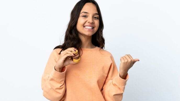 A young woman with long hair holds colorful French macarons and points to the side.