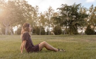 A young woman with long hair sits on a grassy lawn, gazing into the distance with a blurred background.
